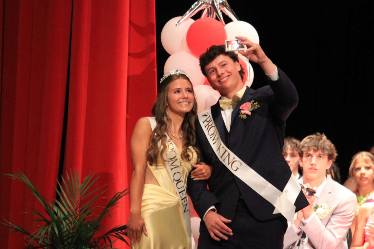Stephanie Novak and Kai Close strike a pose taking a selfie on stage, Novak wearing her crown and the two of them wearing king and queen satchels. The two were announced as class of 2025’s prom king and queen.